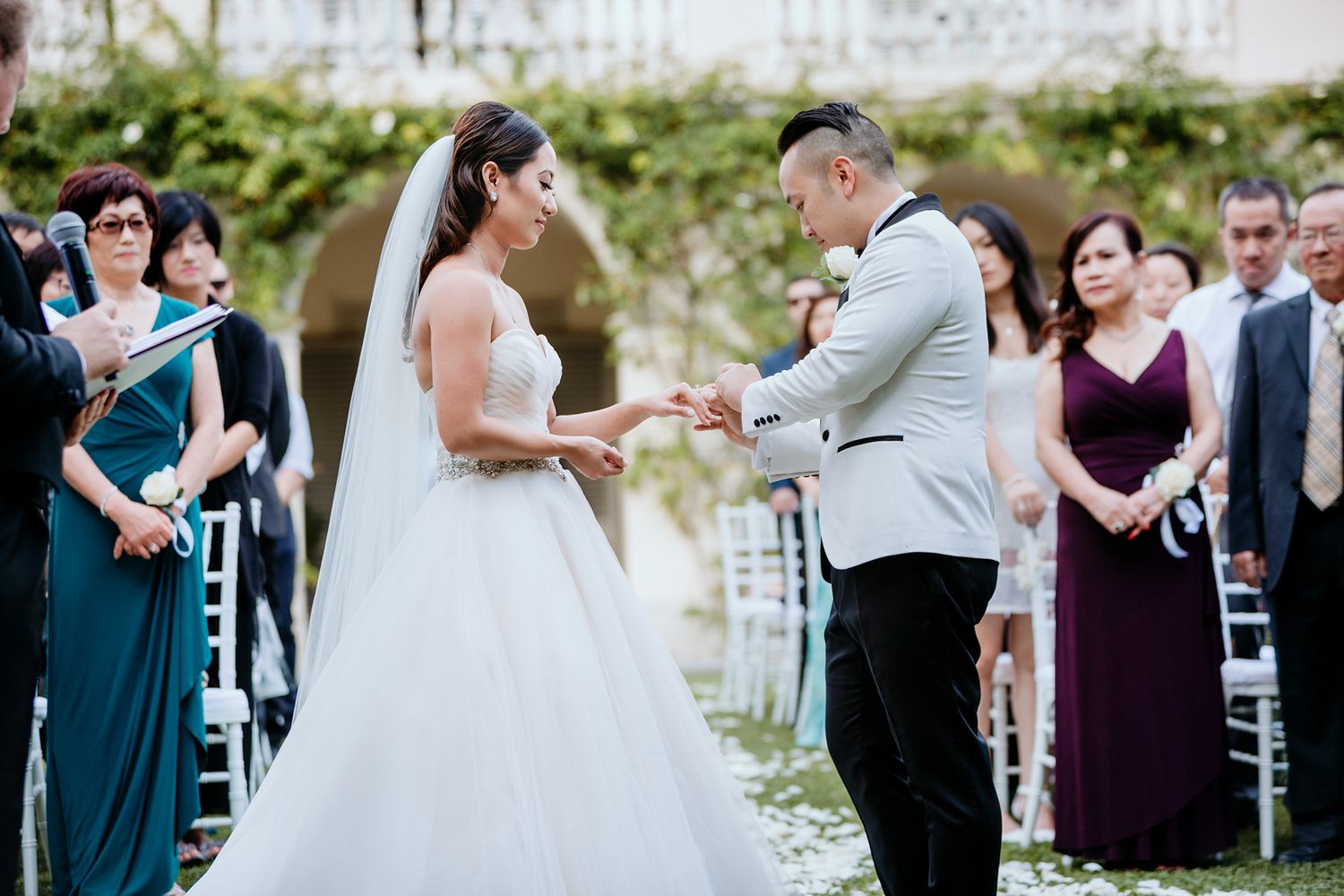 A groom places a wedding ring on his bride's finger at an elegant outdoor garden wedding.