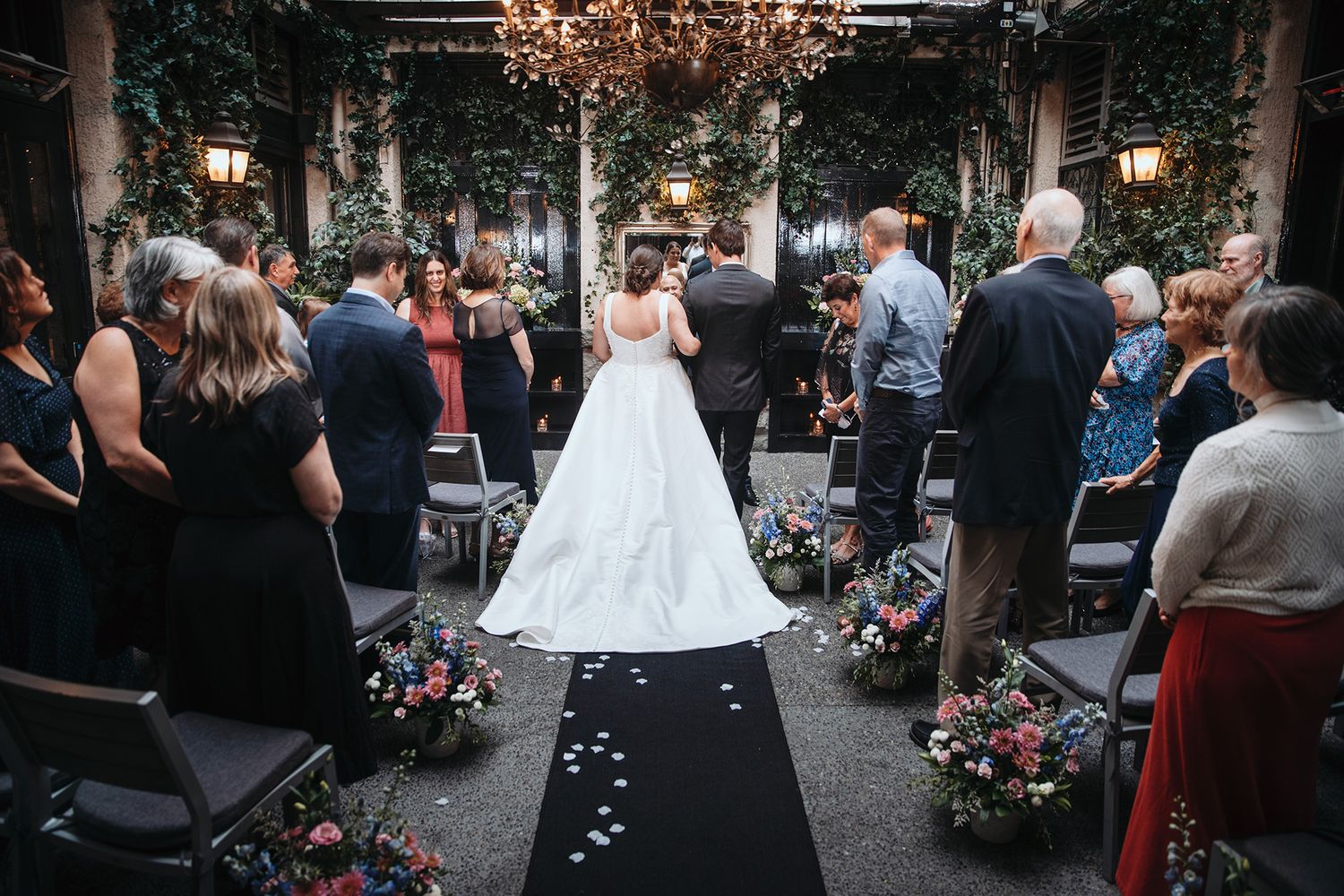 Indoor wedding ceremony under a chandelier with flower petals scattered along a black aisle runner.