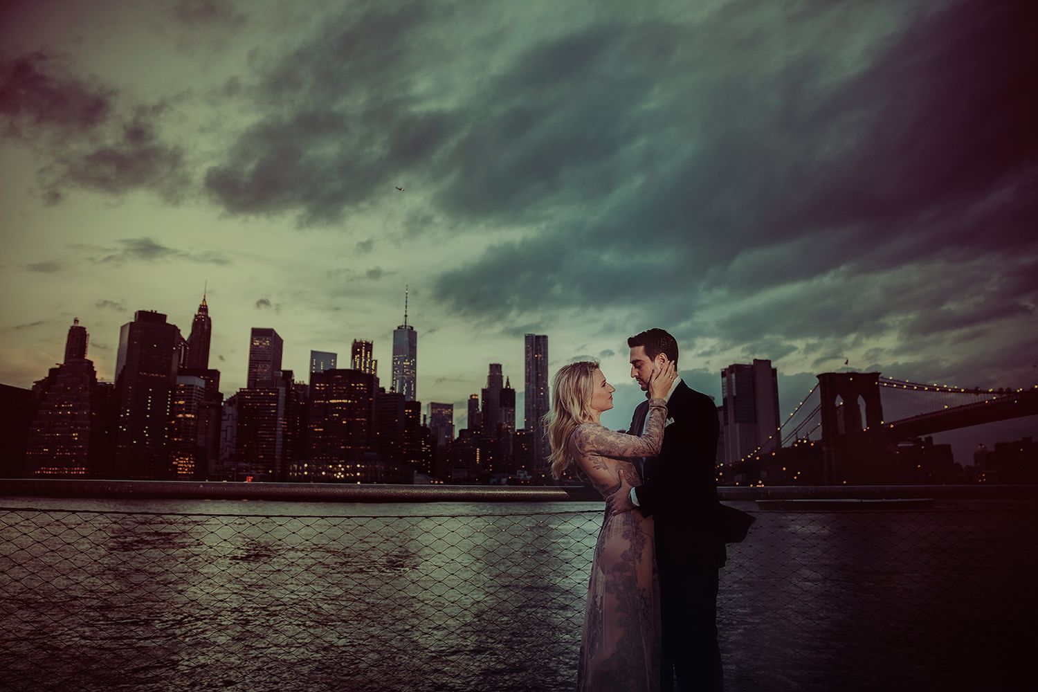 a couple share a romantic moment with the Manhattan skyline silhouetted against dramatic stormy clouds at dusk with Brooklyn Bridge in view.