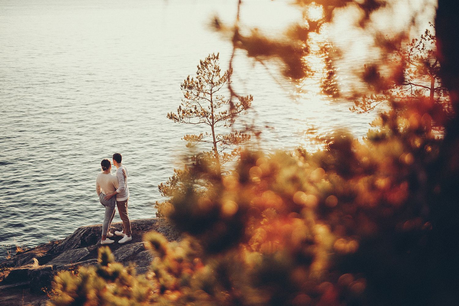 A couple sits together at sunset by the Pacific ocean, surrounded by golden autumn foliage and warm sunlight.