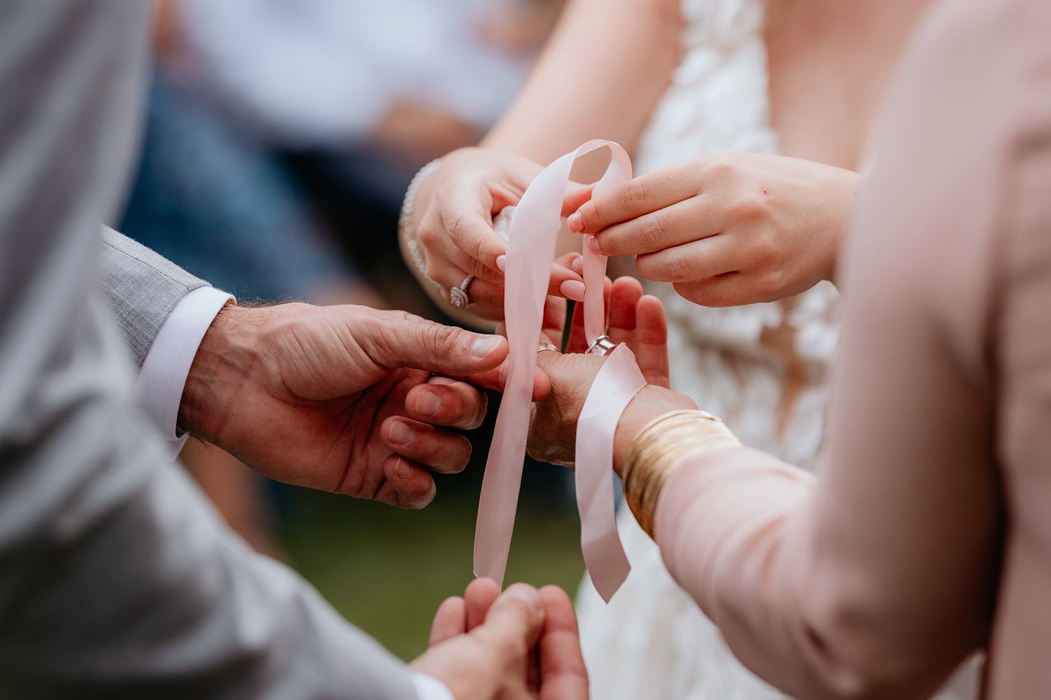 Close-up view of hands holding a delicate pink ribbon during an outdoor wedding ceremony.
