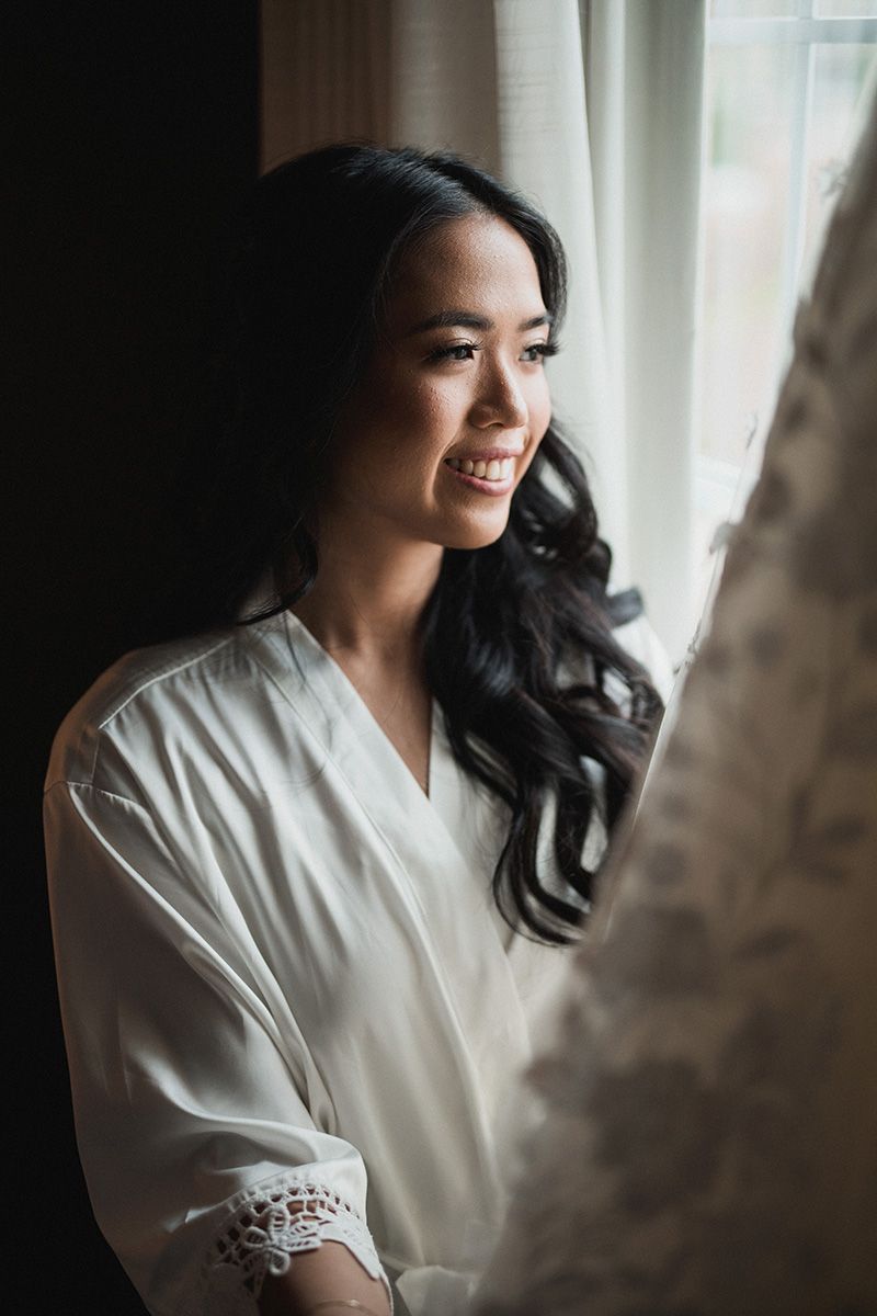 A woman in a white robe smiles warmly by a window with soft natural lighting creating an intimate bridal portrait.