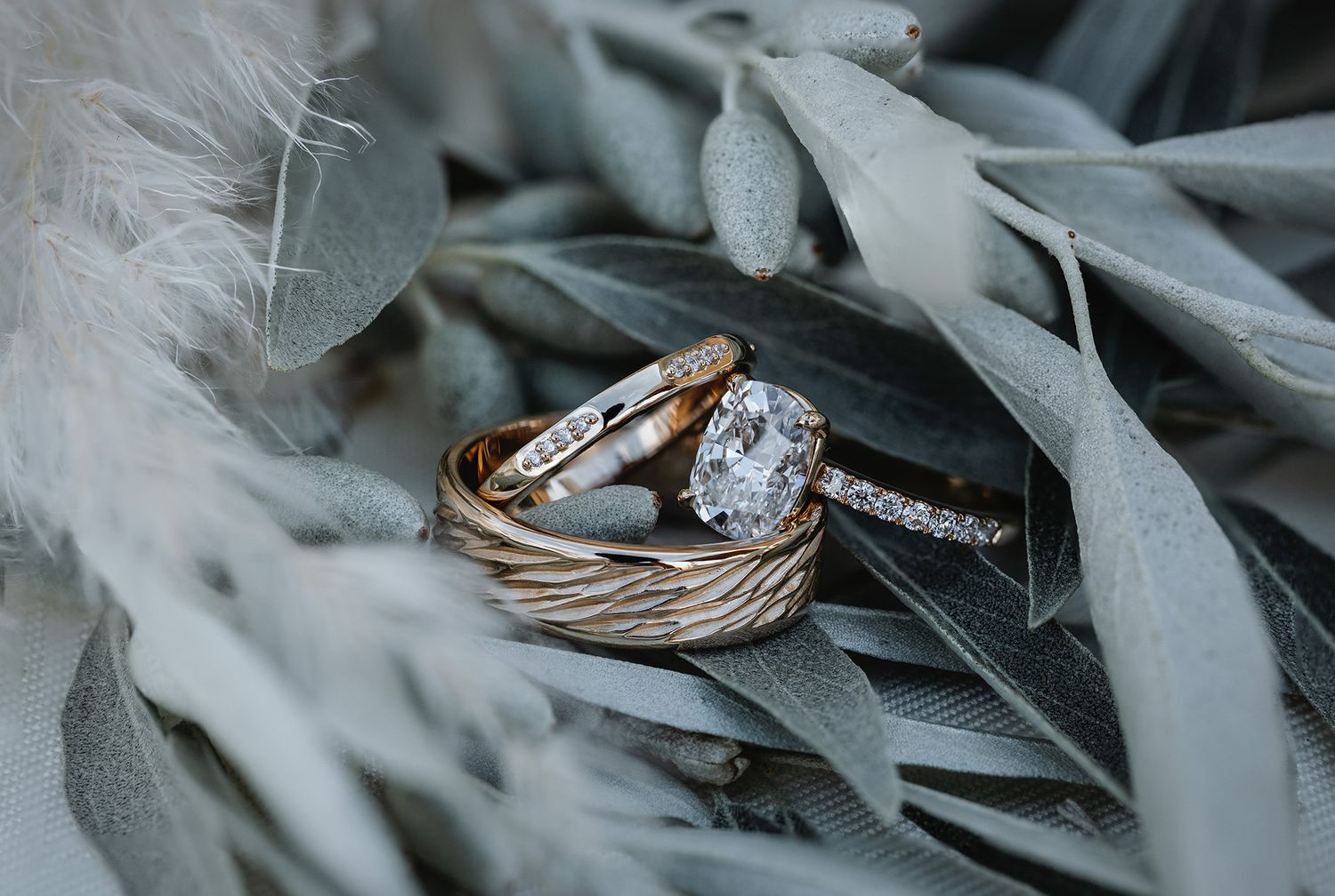 Close-up detail of an elegant engagement ring with diamond solitaire resting on soft grey feathers and leaves.