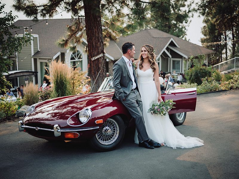 Elegant wedding couple poses with a classic burgundy Jaguar E-Type sports car on a tree-lined residential street.