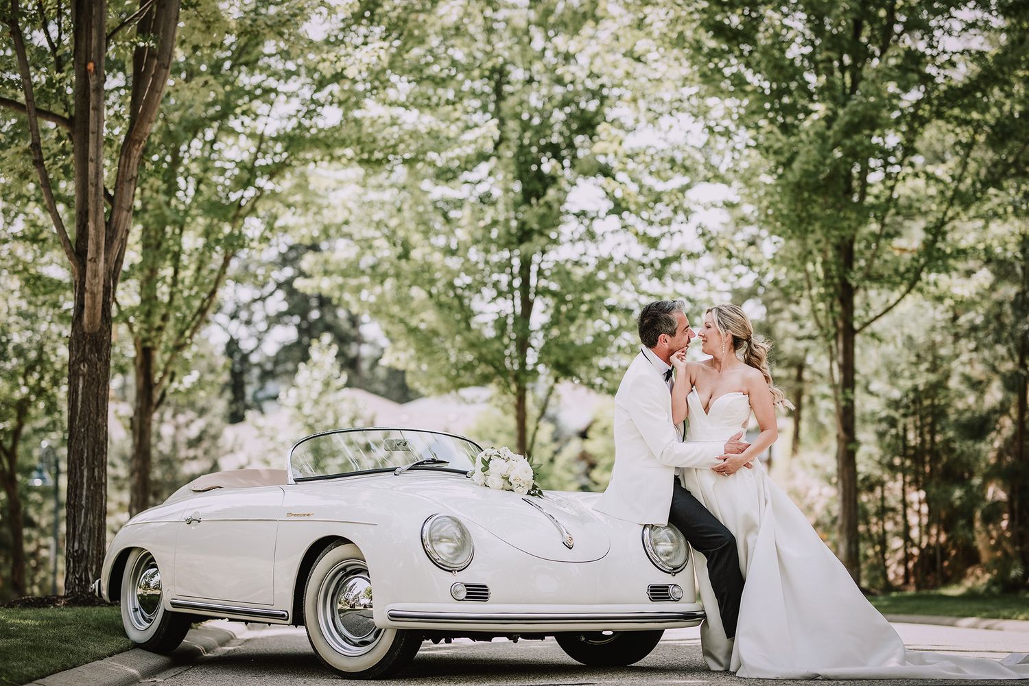 Groom in white suit sits by white vintage Porsche 356 Speedster parked in the shade. His bride leans on him during romantic wedding photos.