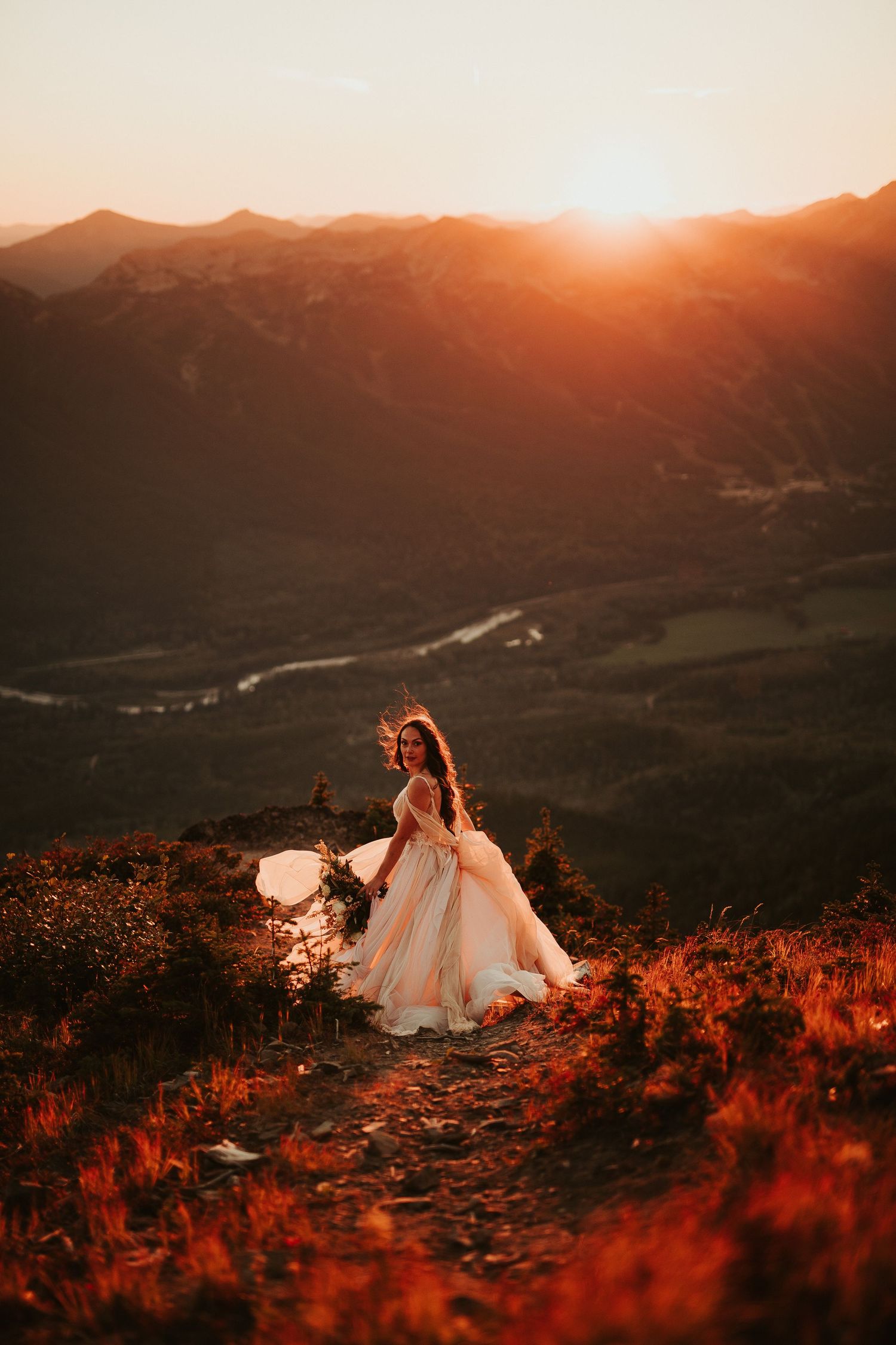 A bride atop a rocky mountain peak at sunset with dramatic orange light illuminating the landscape below.
