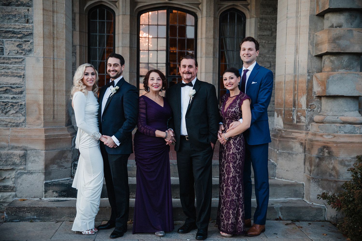 A group of formally dressed people pose together for a wedding photo against a stone building exterior.