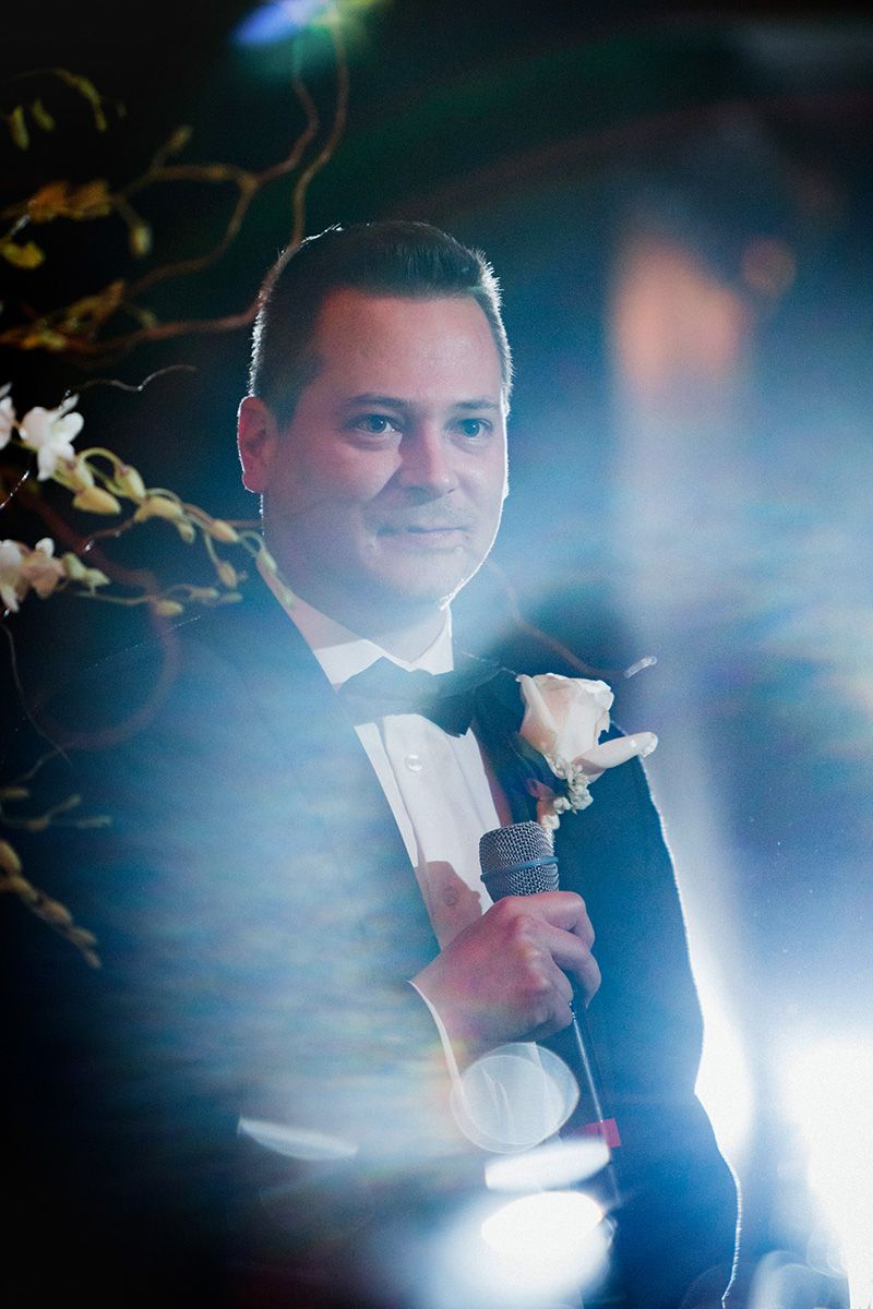 Person in formal black tuxedo and white bowtie holding a microphone while performing in atmospheric lighting.