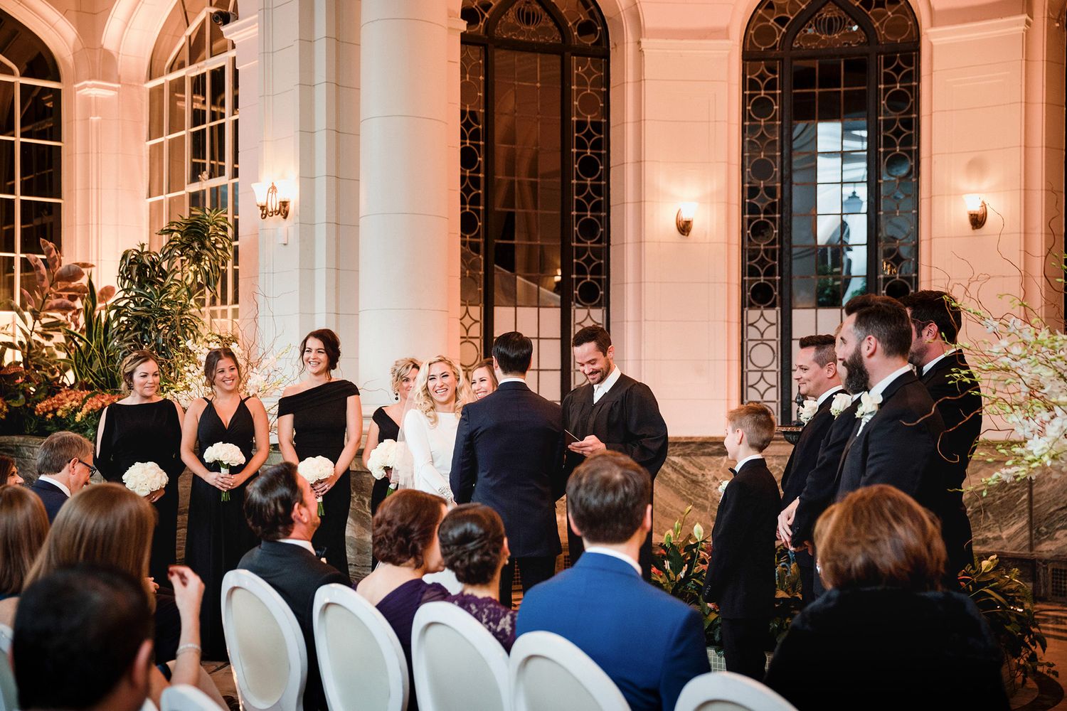 Wedding guests stand in formal attire during an elegant ceremony in a grand ballroom with arched windows and soft lighting.