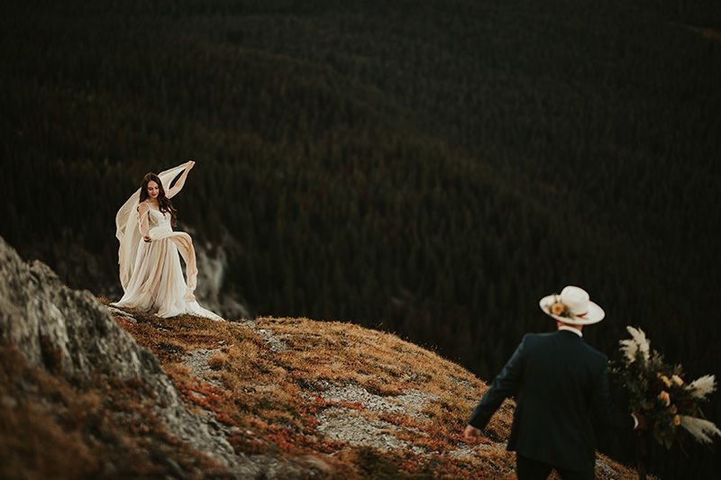 A bride in a flowing white dress stands on a rocky cliff edge against a dramatic dark sky.