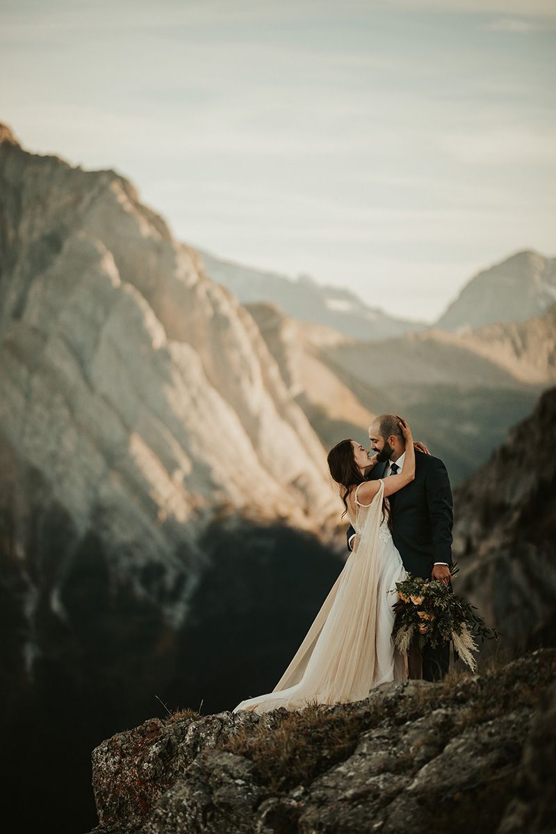 A couple in wedding attire embrace while standing on a rocky mountain ledge with dramatic peaks in the background at sunset.
