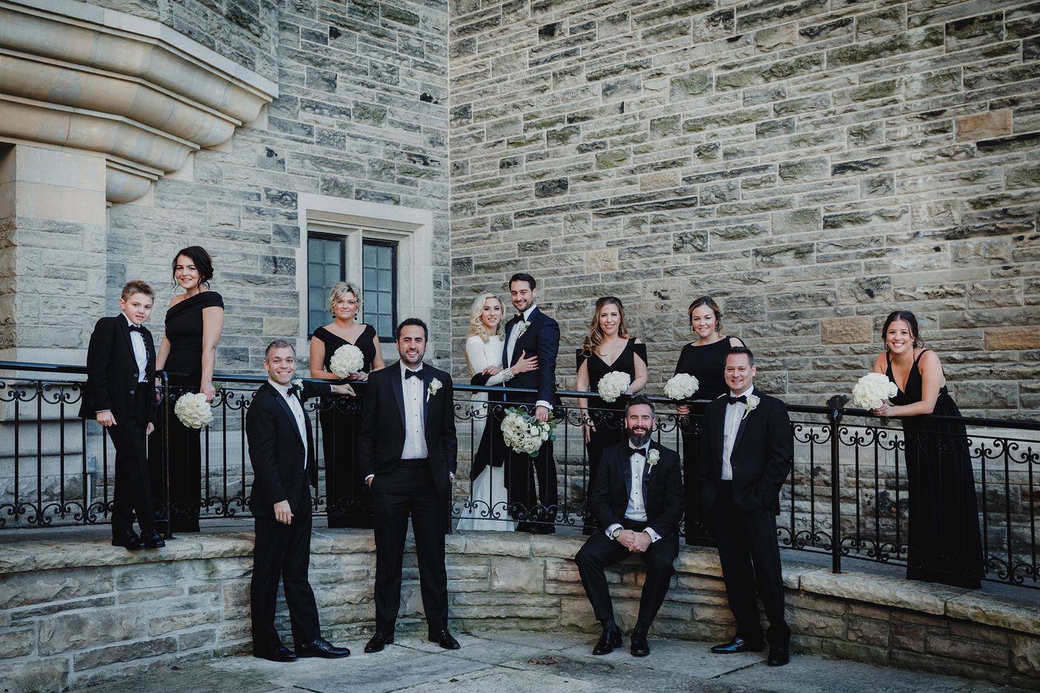 A wedding party in formal attire poses together against a stone wall exterior of a building.