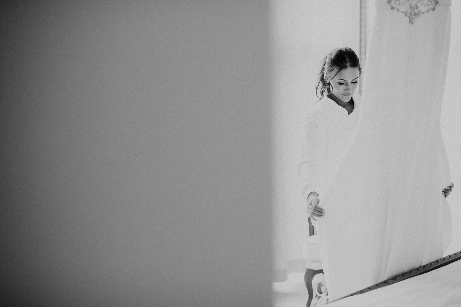 A bride in a robe pulls at the bottom of her wedding dress that hangs from by a window in this black and white photo.