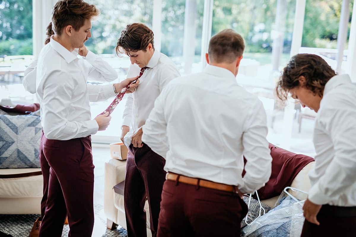 A group of men in white dress shirts and burgundy pants gather in a bright room to help each other with their neck-ties.