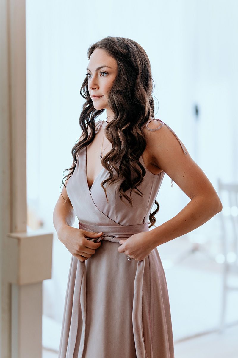 Woman in beige wrap dress poses in natural window light against white interior.