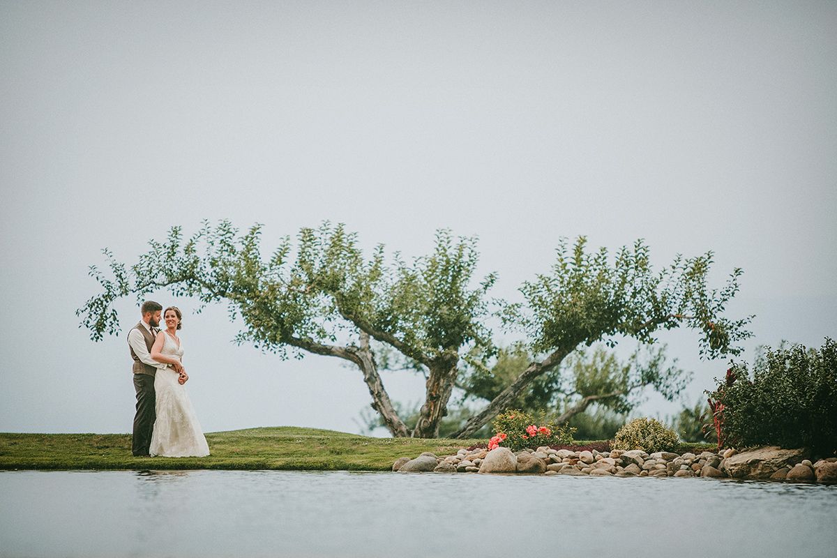 A wedding couple pose beyond a pond with a tree and smokey background.