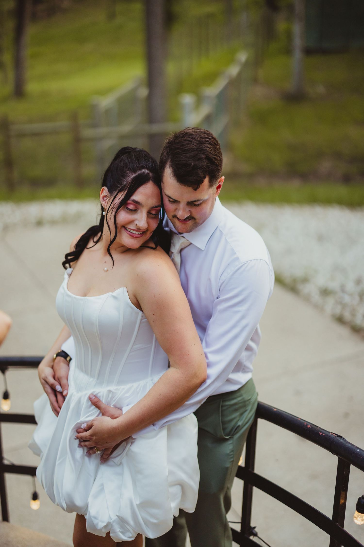 A bride and groom share a tender embrace on outdoor steps, she smiles in a white mini dress as he holds her close.
