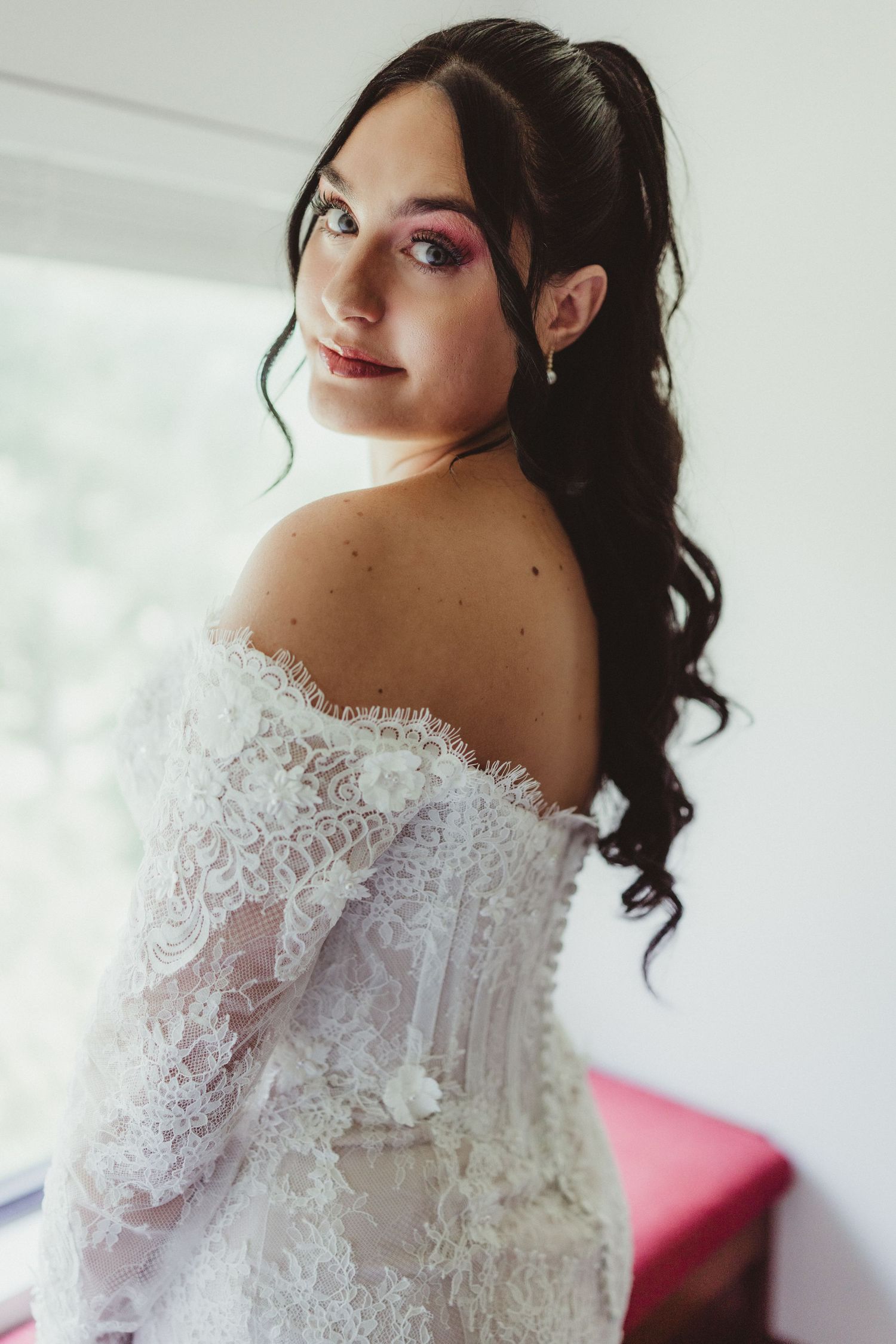 Bride in off-shoulder lace wedding gown with pink eye makeup and dark curly hair, posing elegantly by a window.