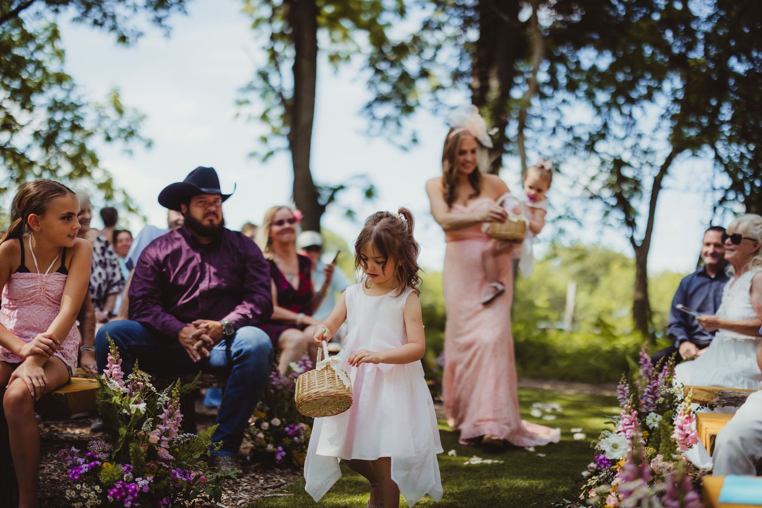 A flower girl in a white dress walks down the aisle at an outdoor wedding ceremony surrounded by guests.