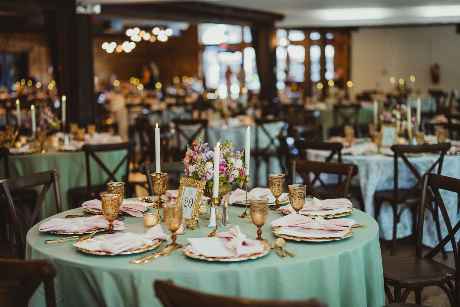 Elegant wedding reception table with mint green linen, gold candles, floral centerpiece, and ornate place settings in a warmly lit venue.