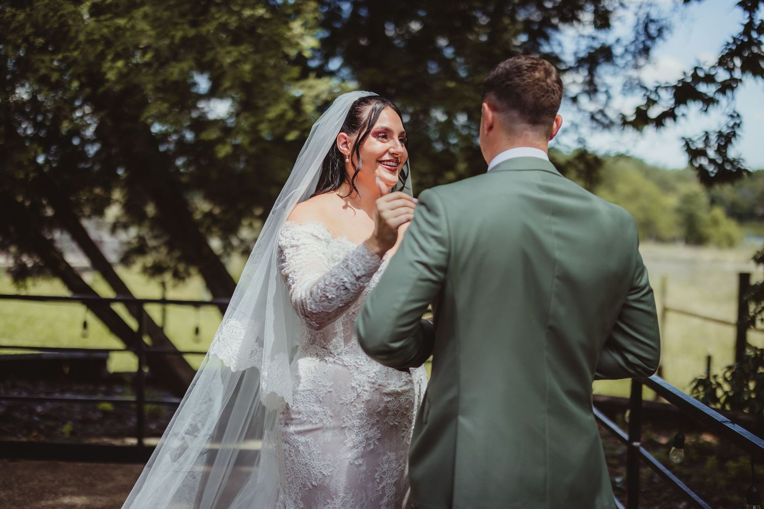 A bride in a lace gown and veil laughs joyfully as a groom in a sage suit gently touches her face outdoors.