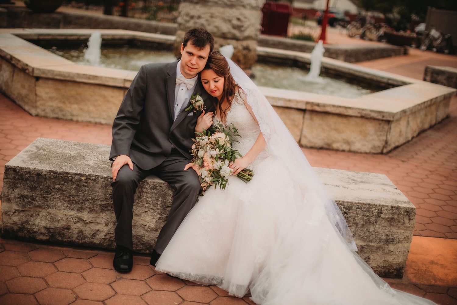 Newlyweds share tender moment while seated on stone steps by fountain.