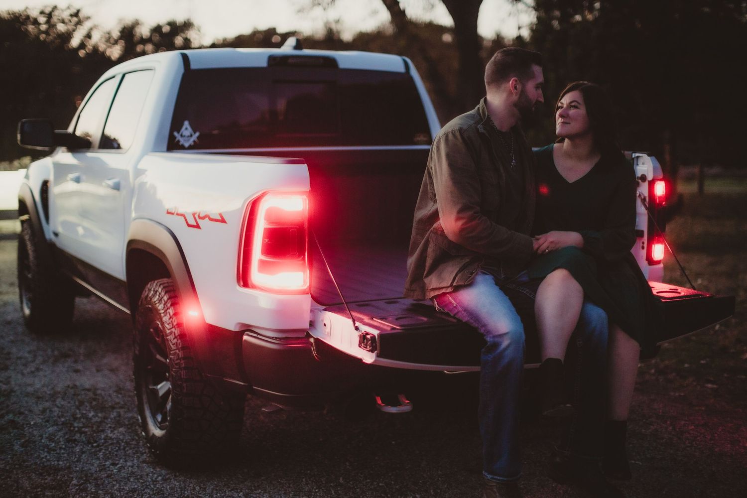Modern pickup truck with illuminated taillights as couple sits on tailgate at dusk.