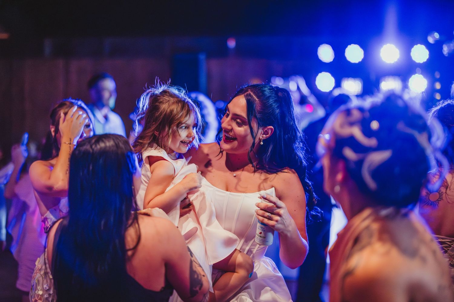 Bride in white dress dancing joyfully with guests at a vibrant wedding reception under colorful lights.