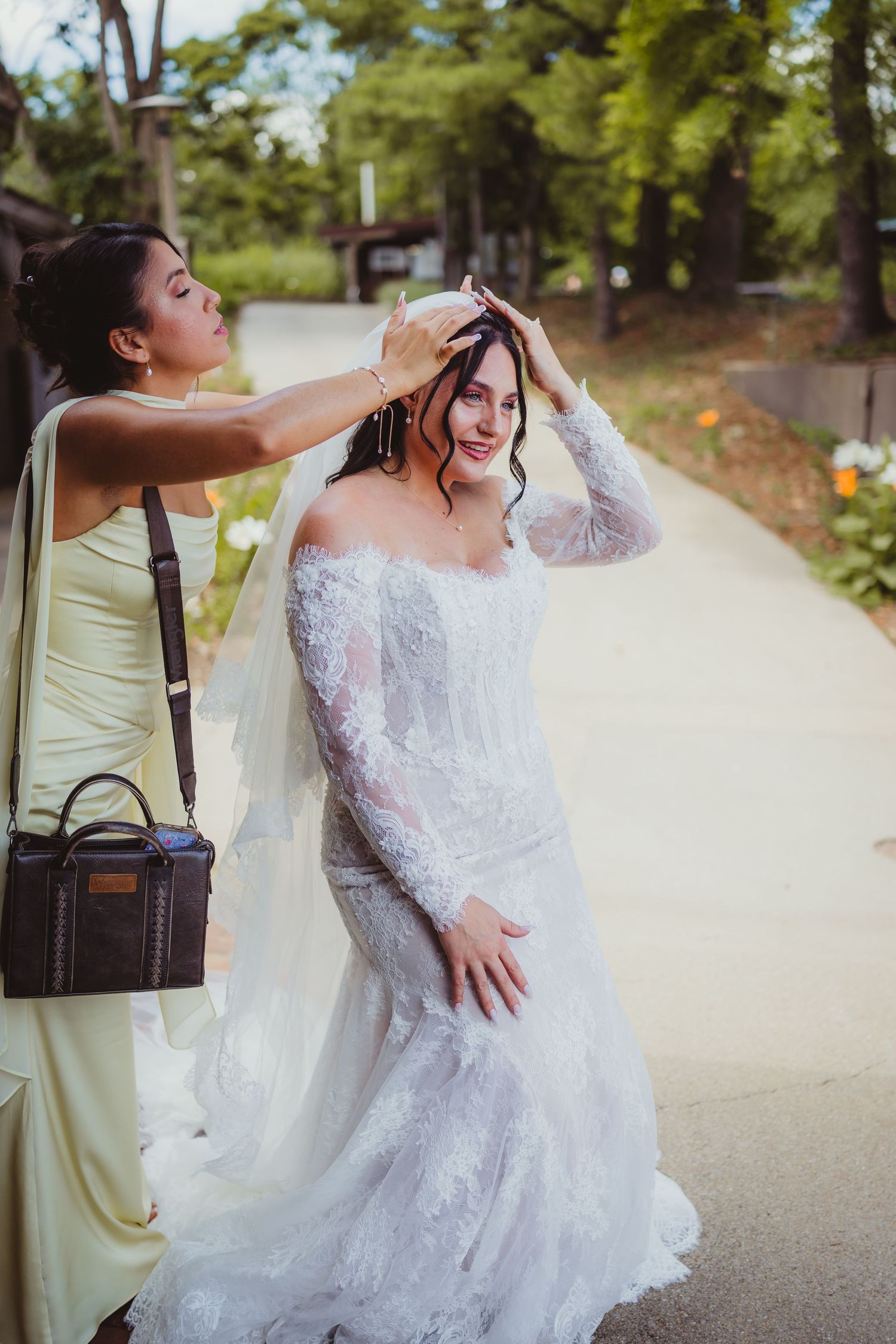 Bridesmaid adjusting bride's veil in white lace off-shoulder gown outdoors on a sunny wedding day.