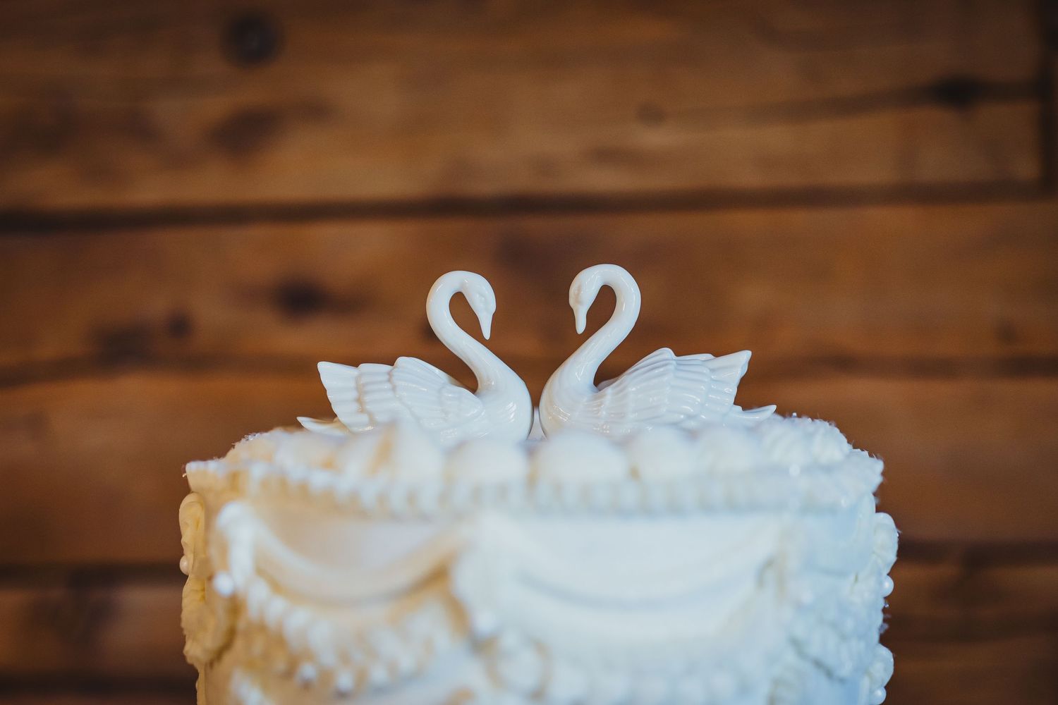 White wedding cake topped with two elegant swan figurines facing each other, set against a rustic wood background.