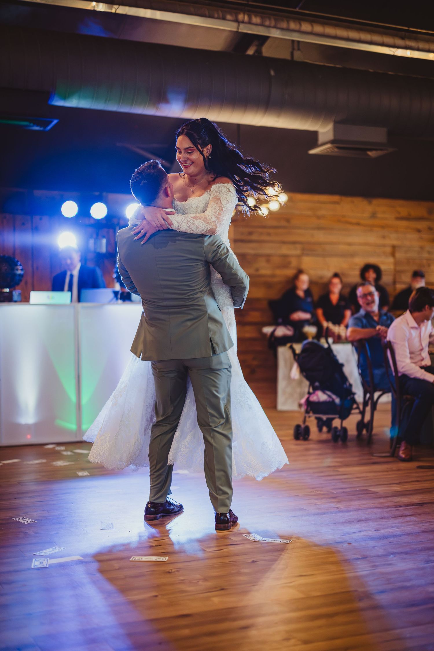 Groom lifts bride during first dance at wedding reception with guests watching in background.
