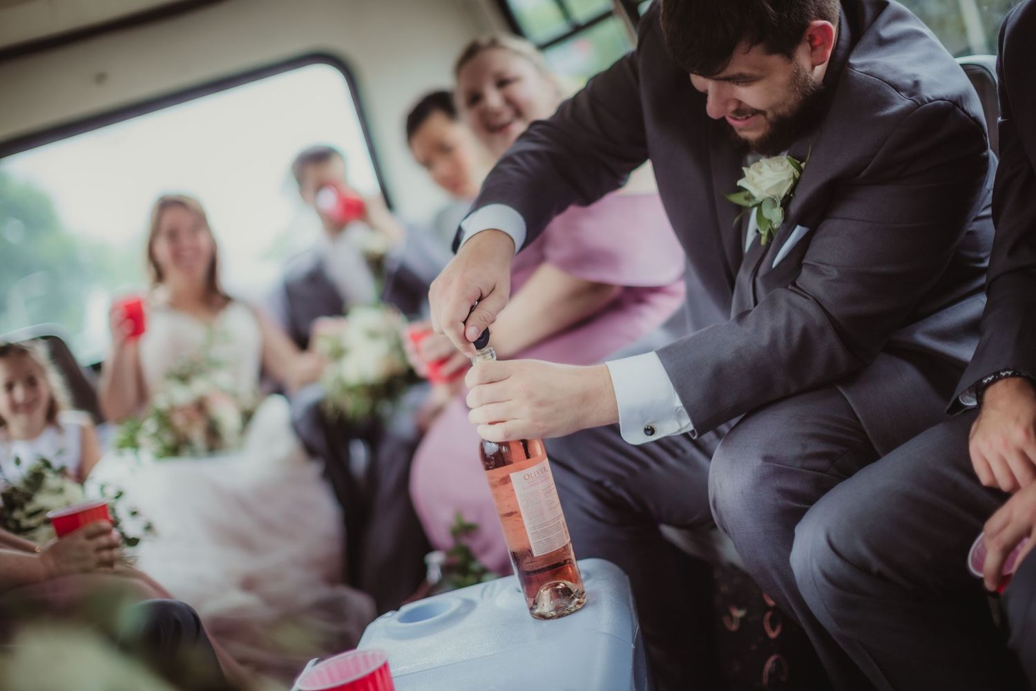 Person in gray suit pours drinks for wedding party members on a shuttle bus.