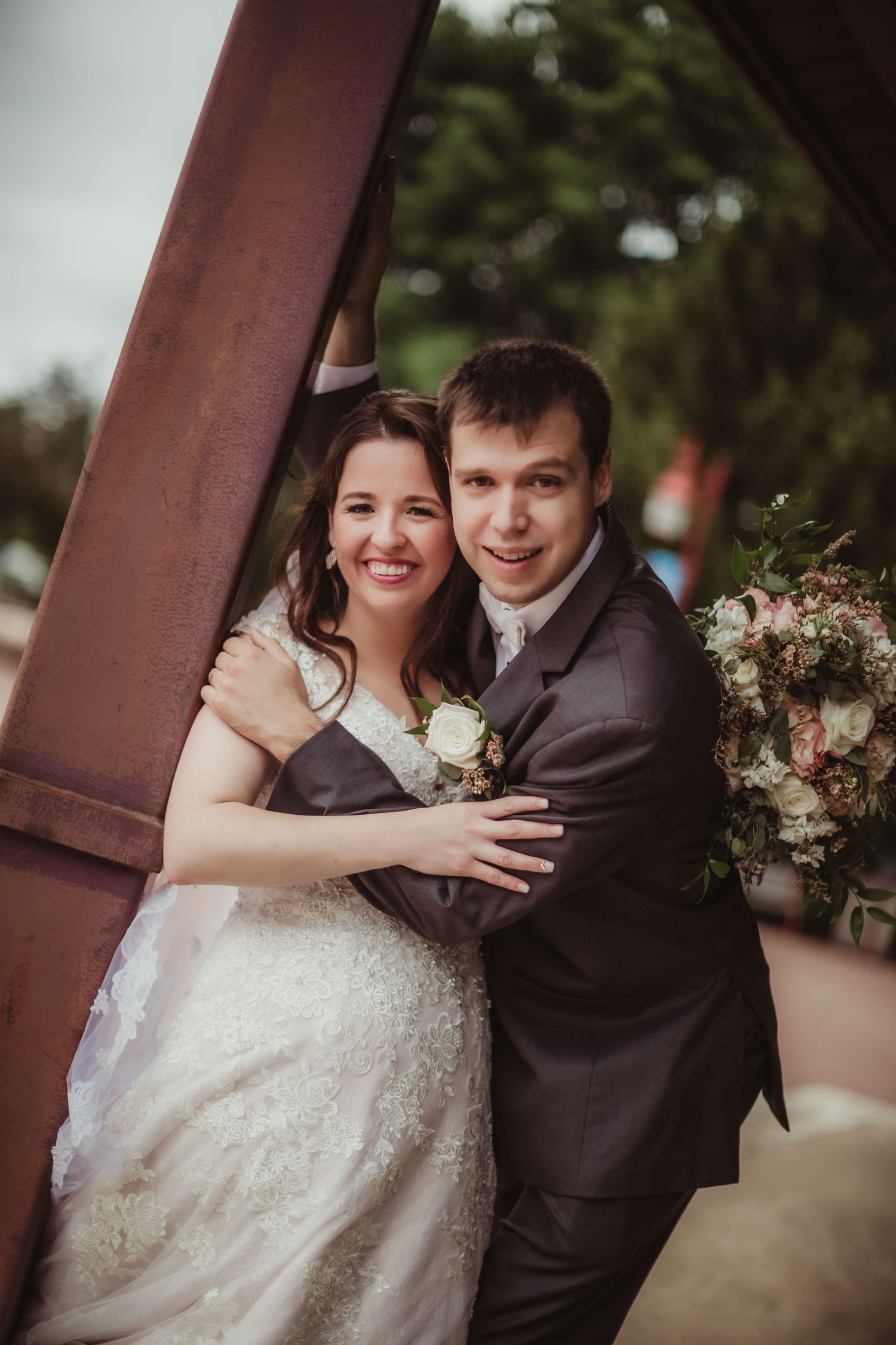 A joyful wedding couple shares a tender moment while posing beside a metal column outdoors.