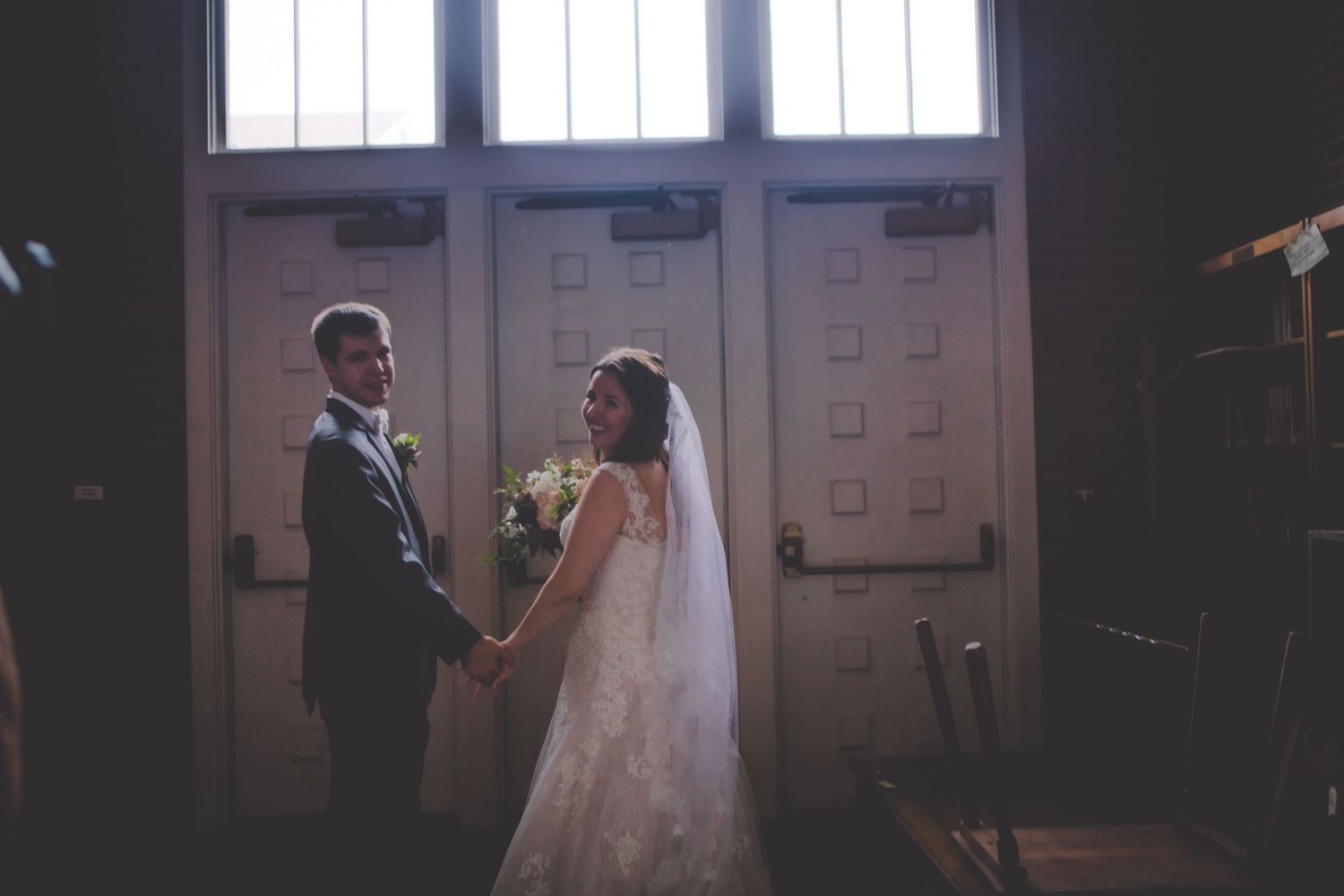 Newlyweds share an intimate moment before church windows in dramatic lighting.