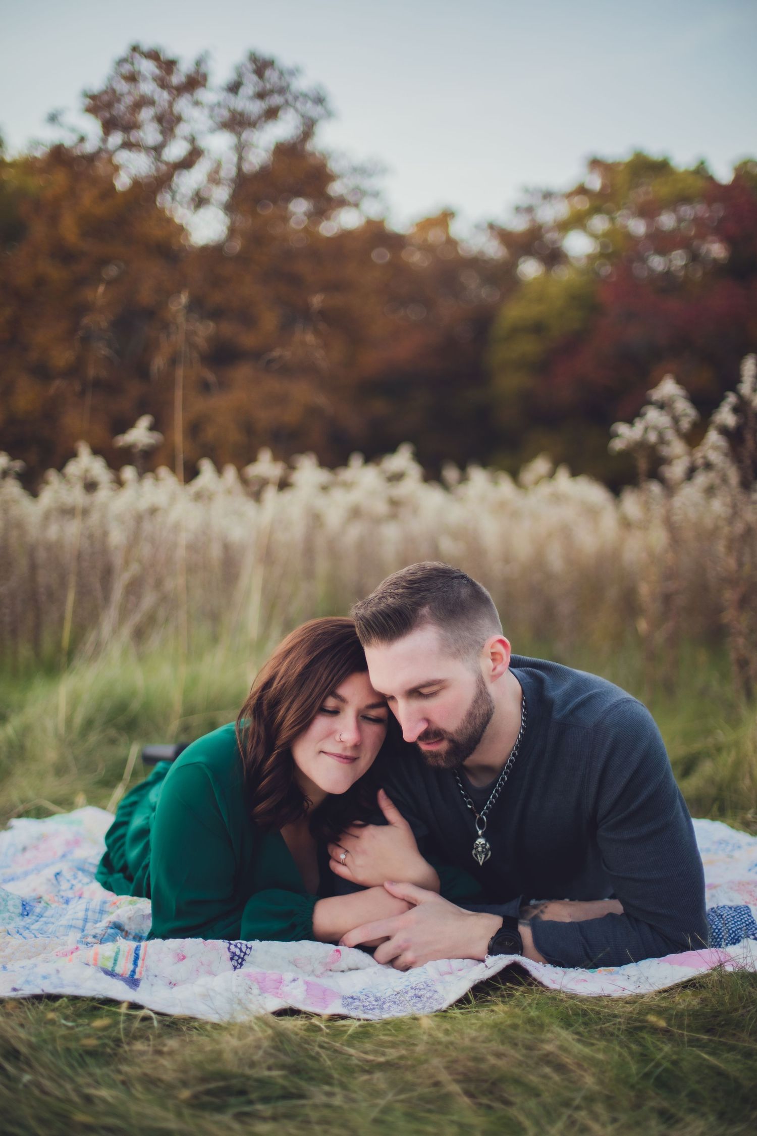 Romantic autumn engagement photo of couple cuddling on blanket in grass field.