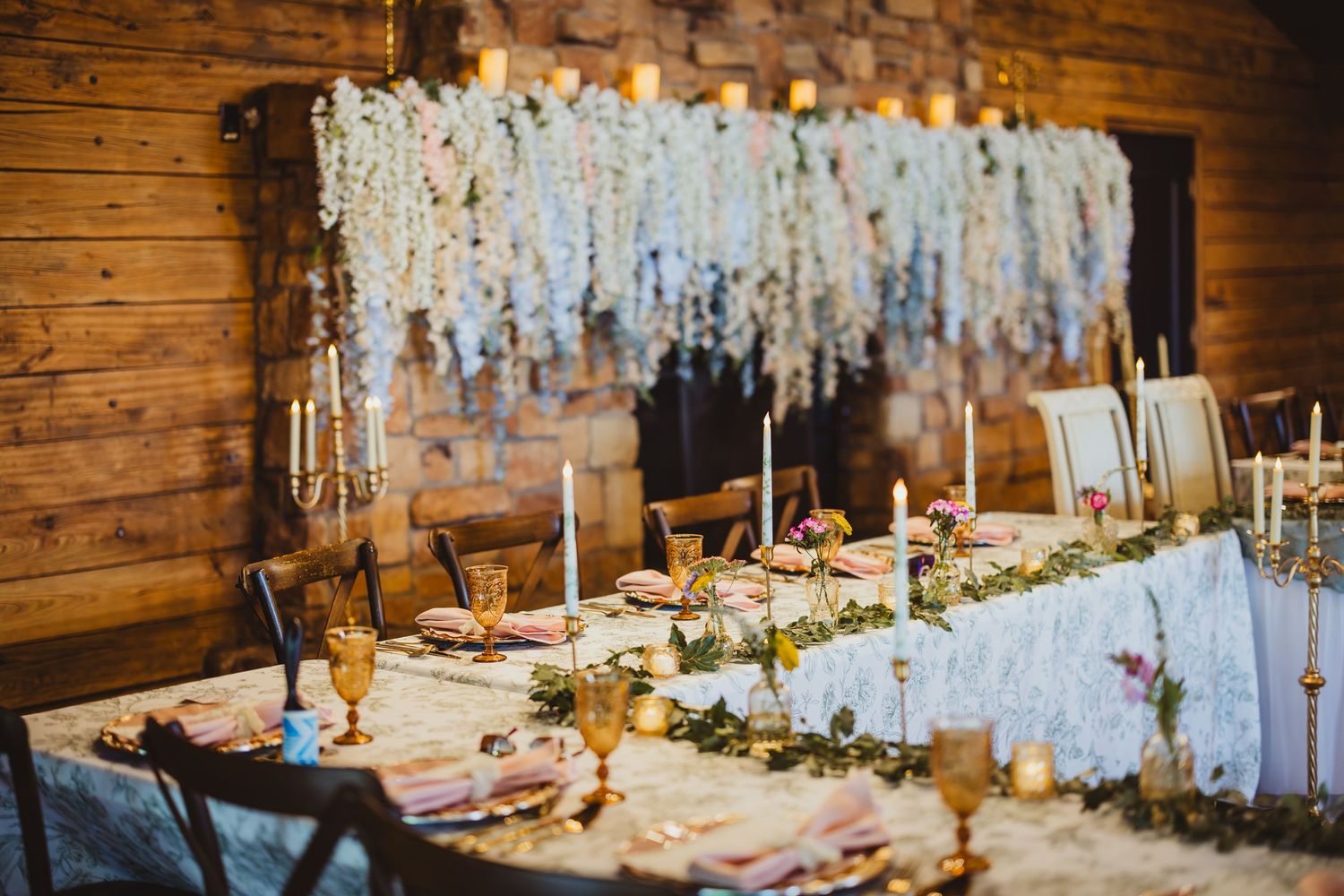 Elegant wedding reception table with white floral backdrop, candles, greenery runner, and gold accents in a rustic barn venue.