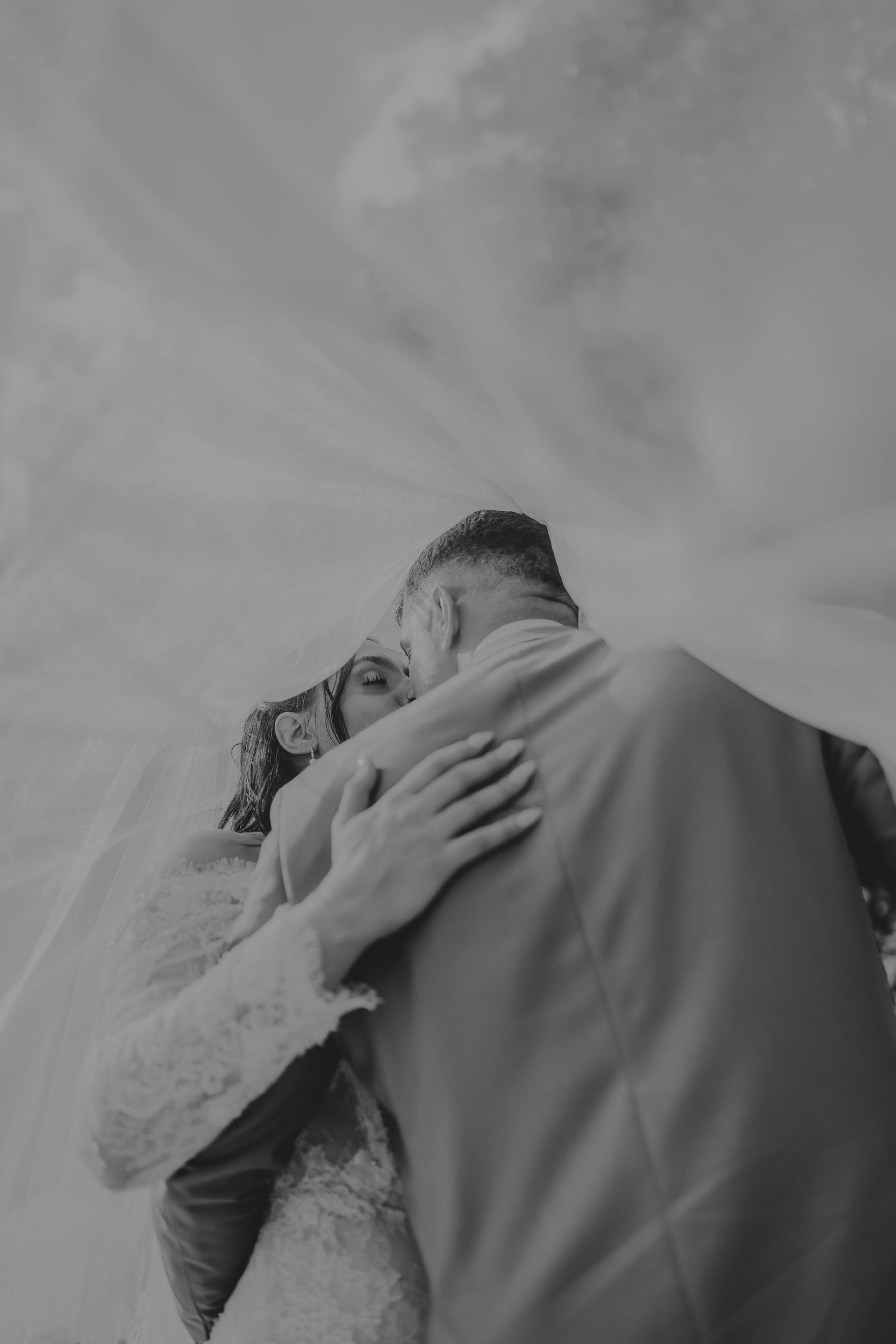 Black and white photo of bride and groom embracing tenderly under a flowing veil on their wedding day.