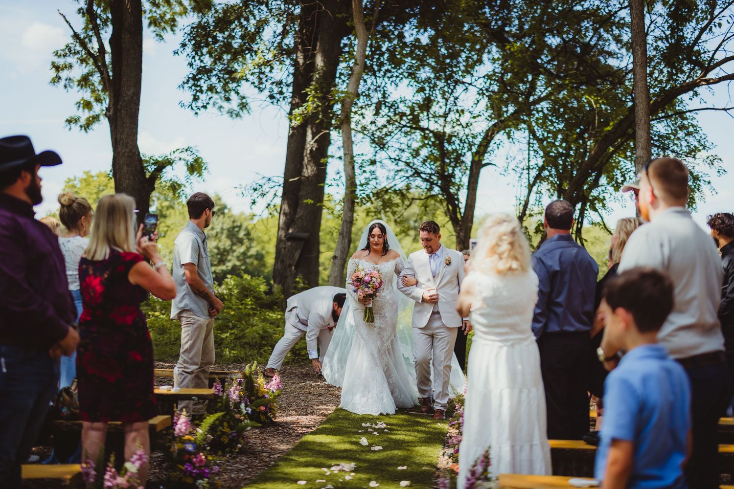 Outdoor wedding ceremony in a wooded garden setting with bride, groom, and guests gathered along a rustic aisle.