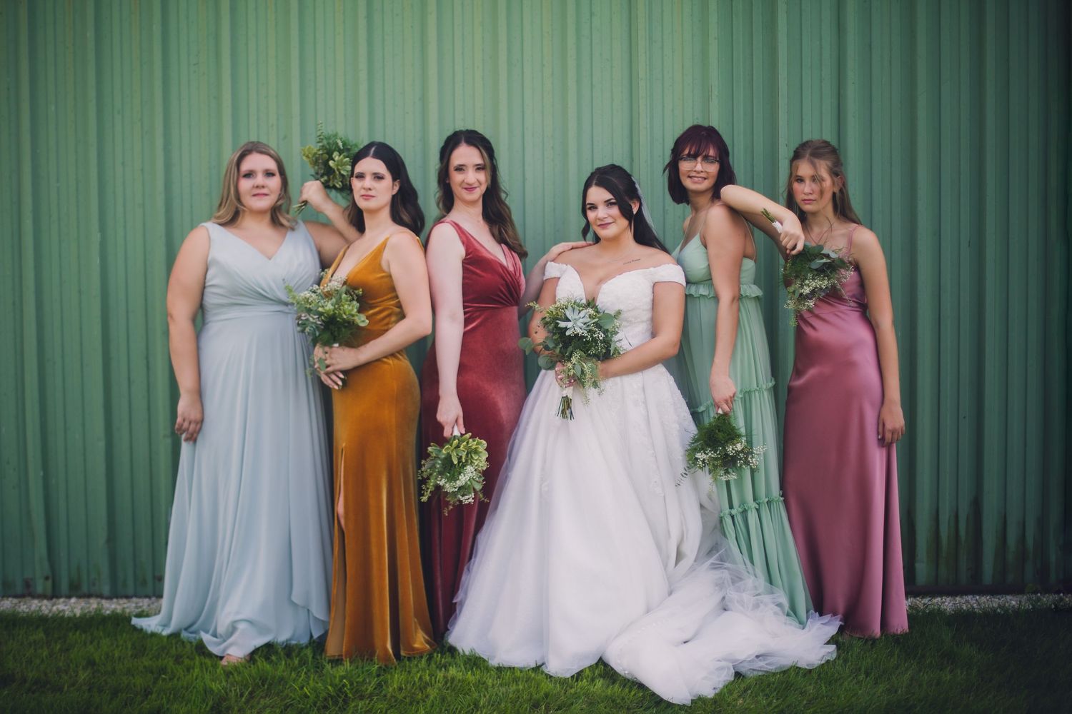 Wedding party in coordinated pastel dresses poses against green corrugated metal backdrop.
