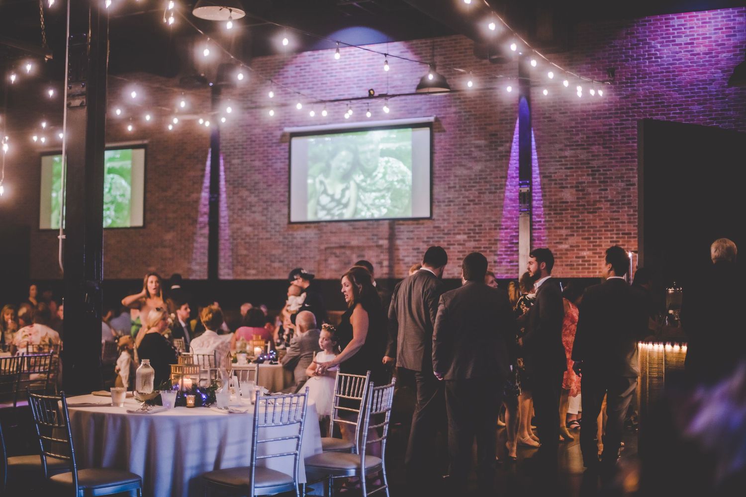 Wedding reception hall with string lights, brick walls and projection screens.