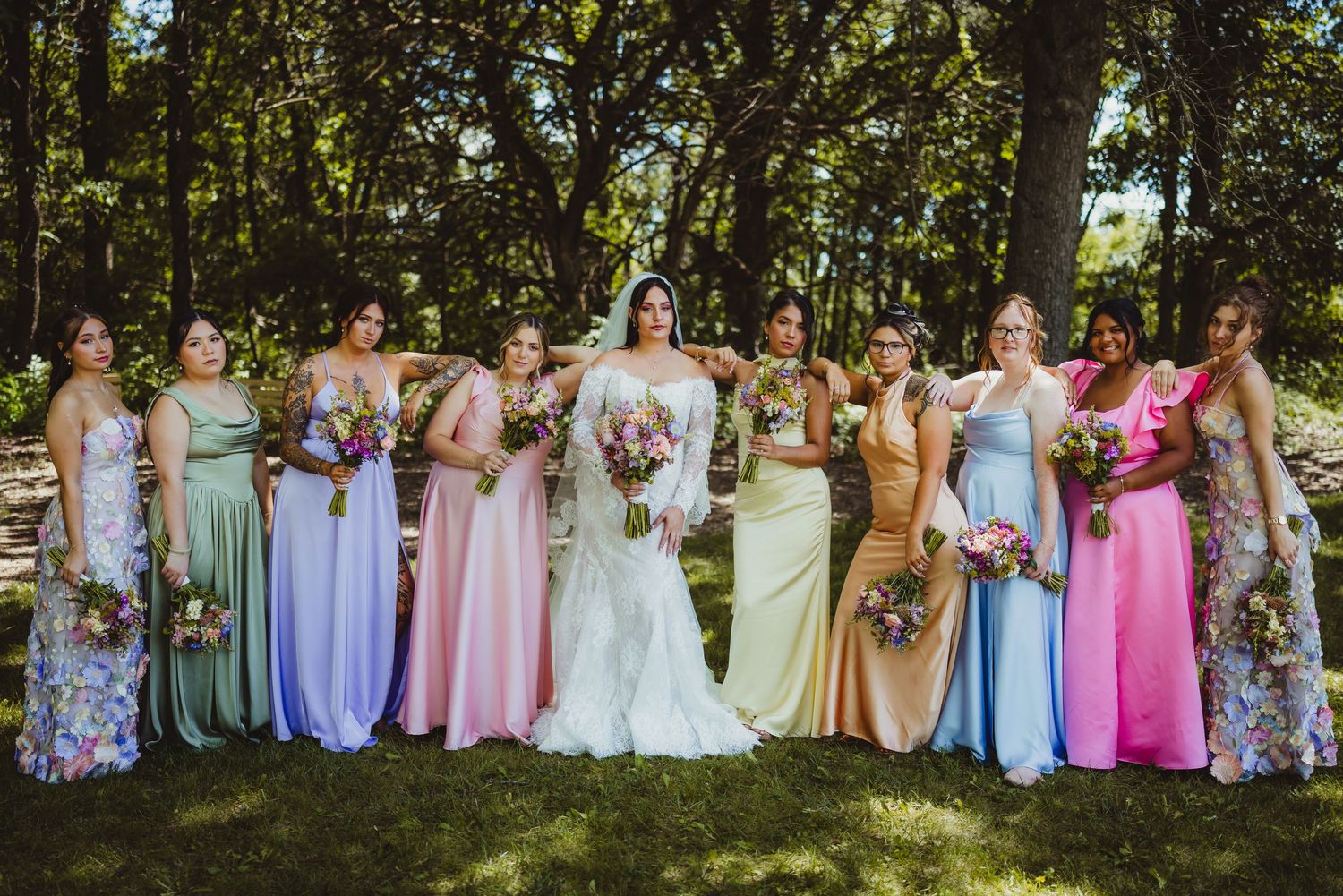 Bride in white gown poses with colorful bridesmaids in rainbow dresses holding bouquets outdoors in a lush green setting.