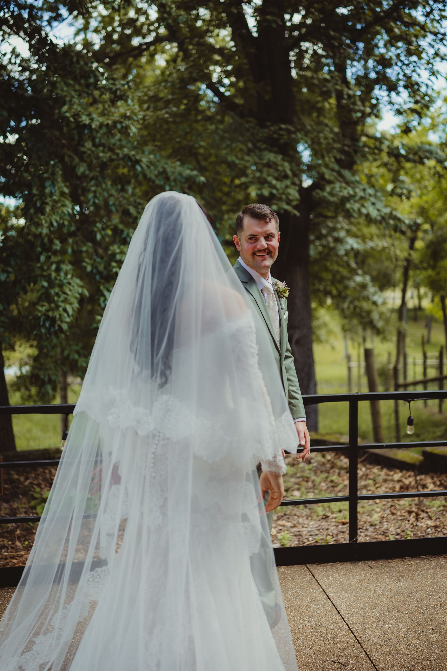 Bride in lace wedding gown and veil holds groom's hand outdoors as he smiles at her in a green suit.