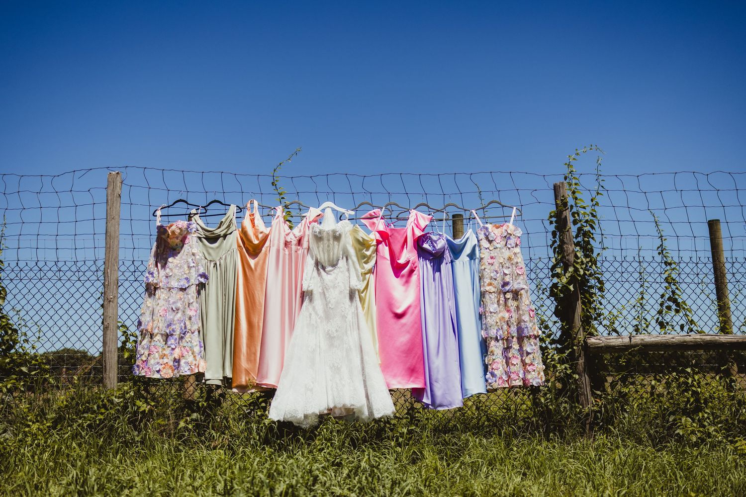 Colorful bridesmaid dresses and white wedding gown hanging on wire fence under bright blue sky outdoors.