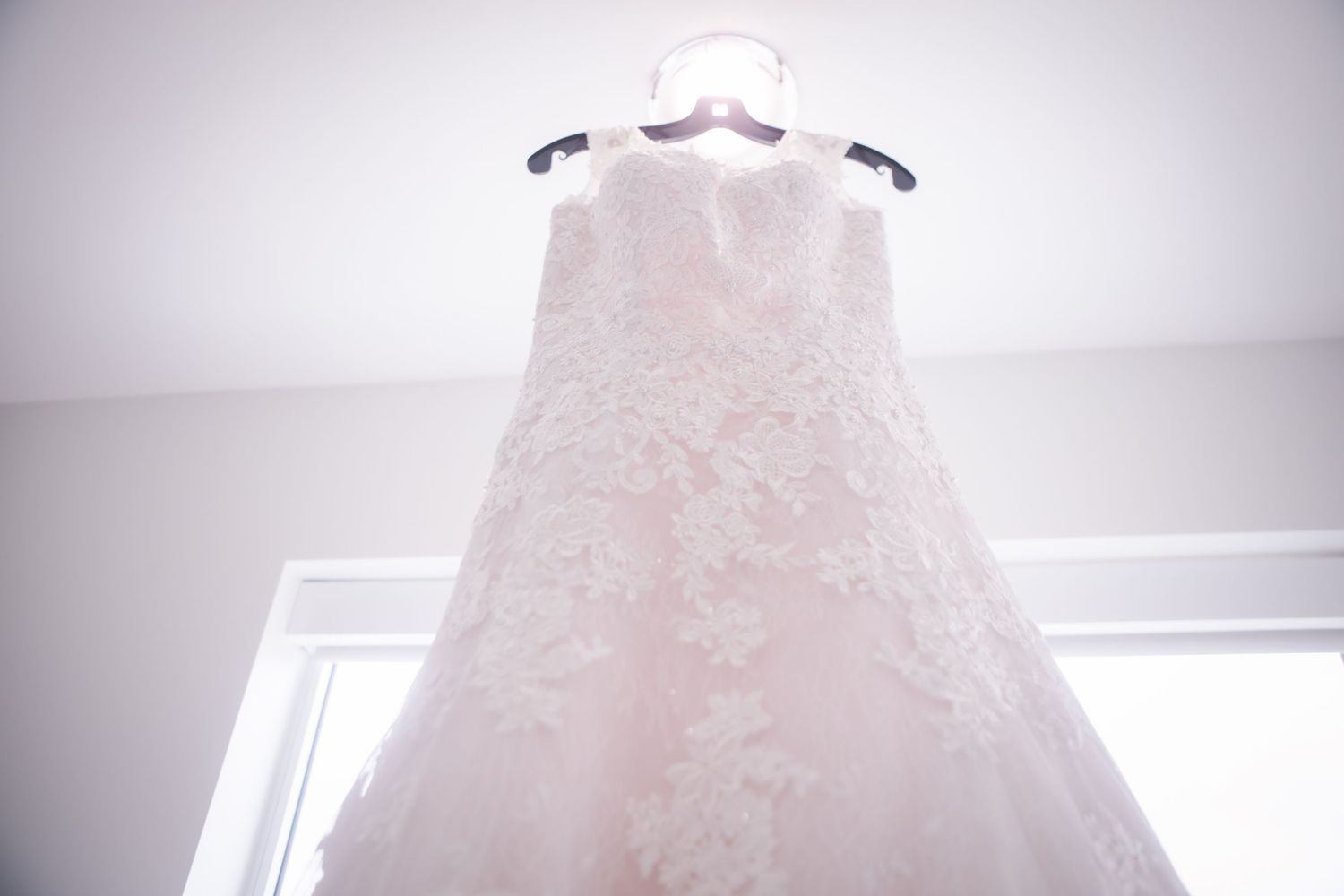 Light pink wedding gown displayed against bright window in minimalist white room.