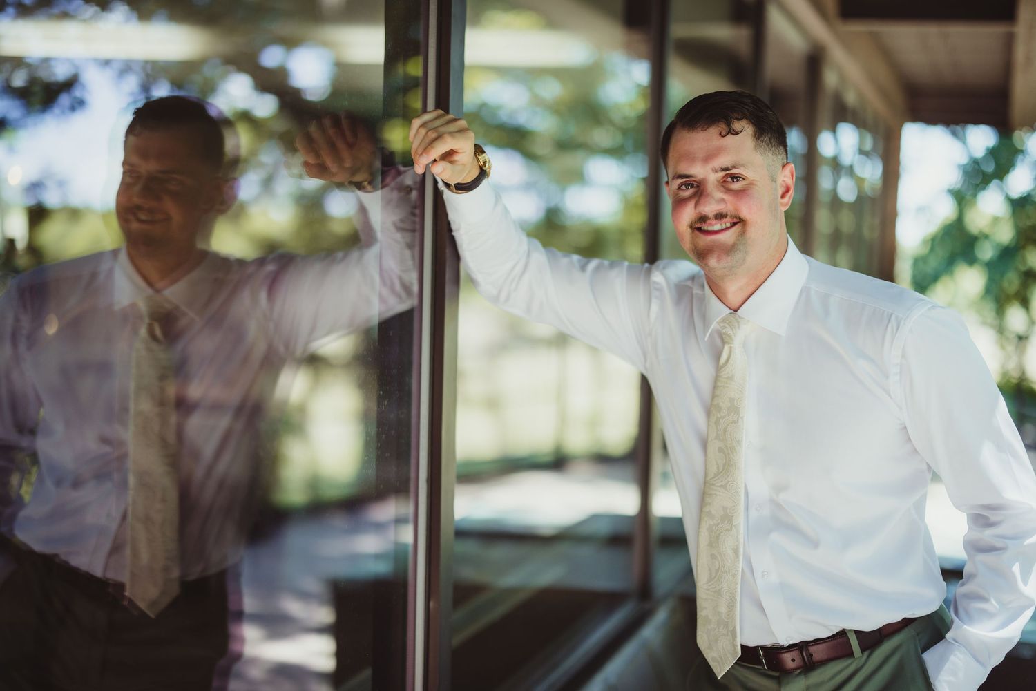 Smiling man in white dress shirt and gold tie leaning against a glass wall with his reflection visible.