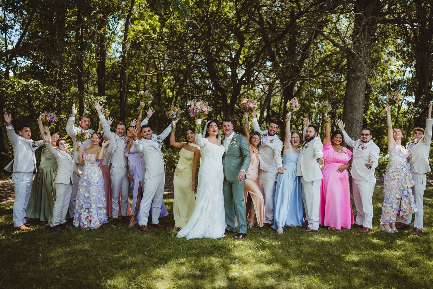Wedding party celebrating outdoors under trees, bride in white gown, groom in sage suit, colorful bridesmaids and groomsmen cheering.