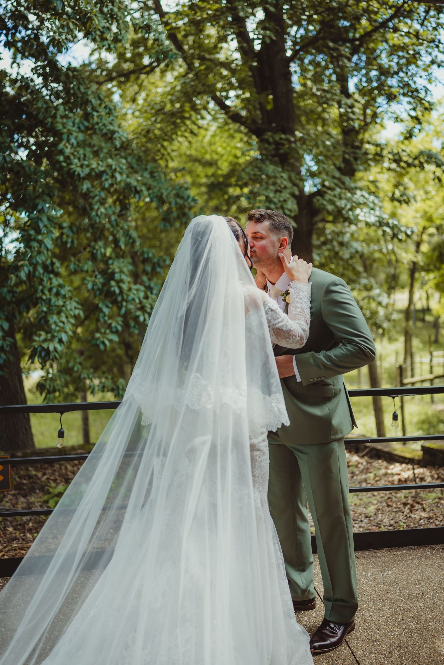 Bride and groom share a romantic kiss outdoors, bride in white gown with long veil, groom in sage green suit.