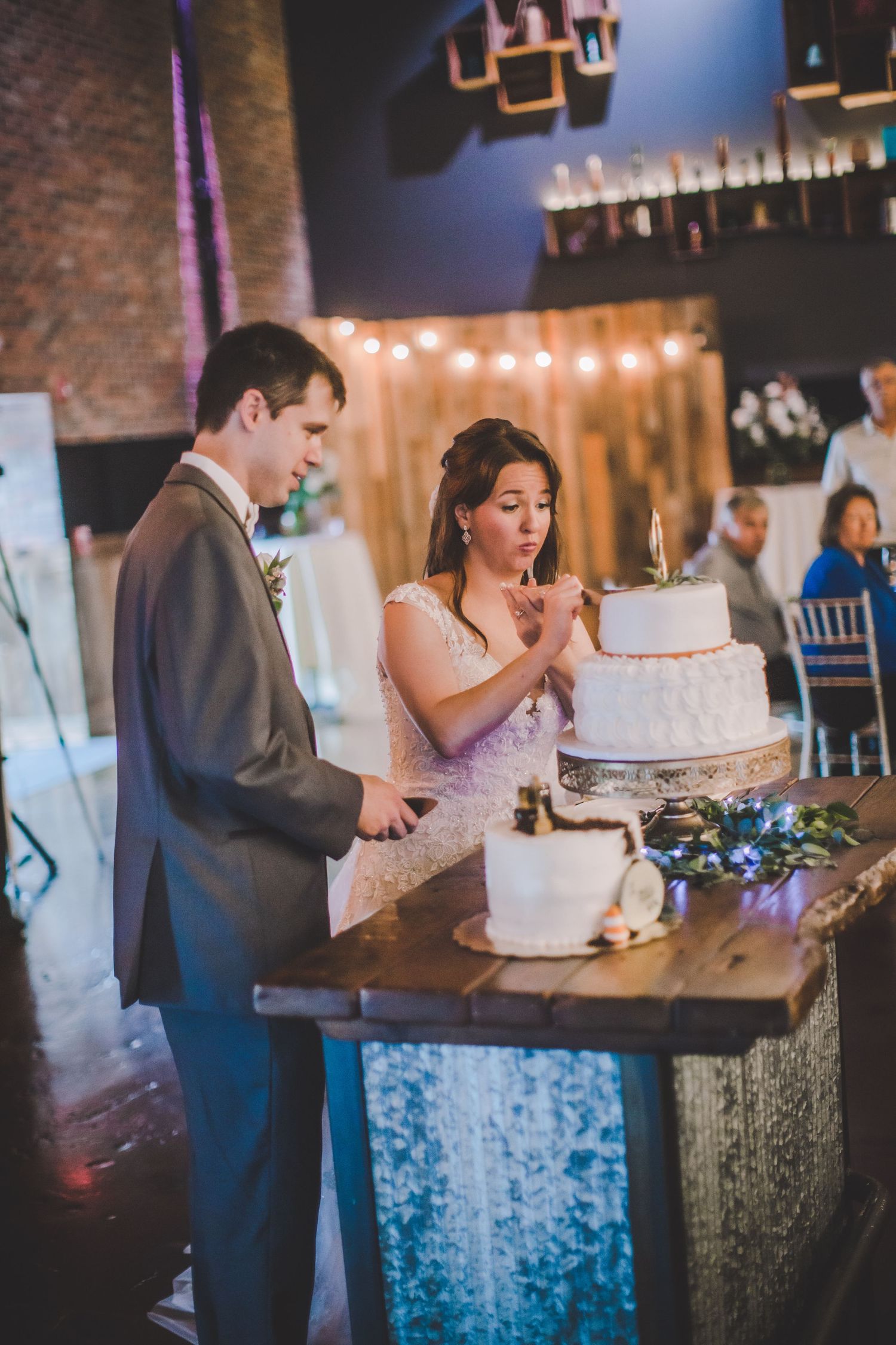 Couple shares cake cutting moment at wedding reception with string lights and brick backdrop.