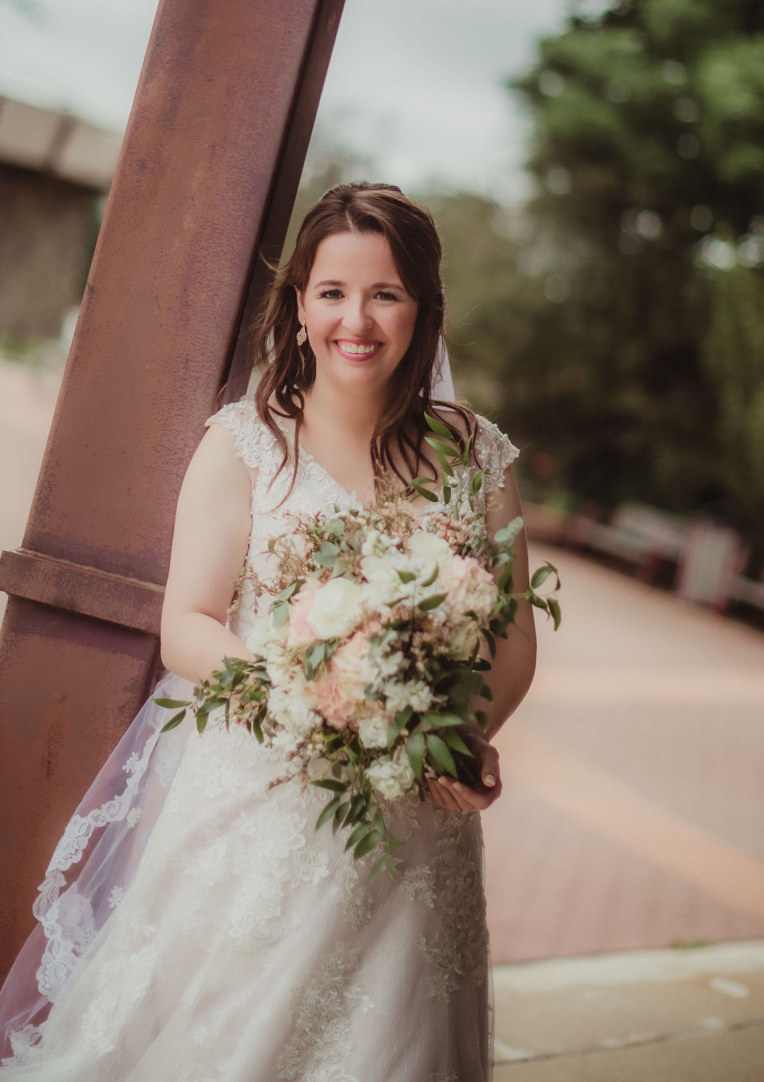 A bride in a lace wedding gown holds a soft pastel bouquet while posing by a metal column.