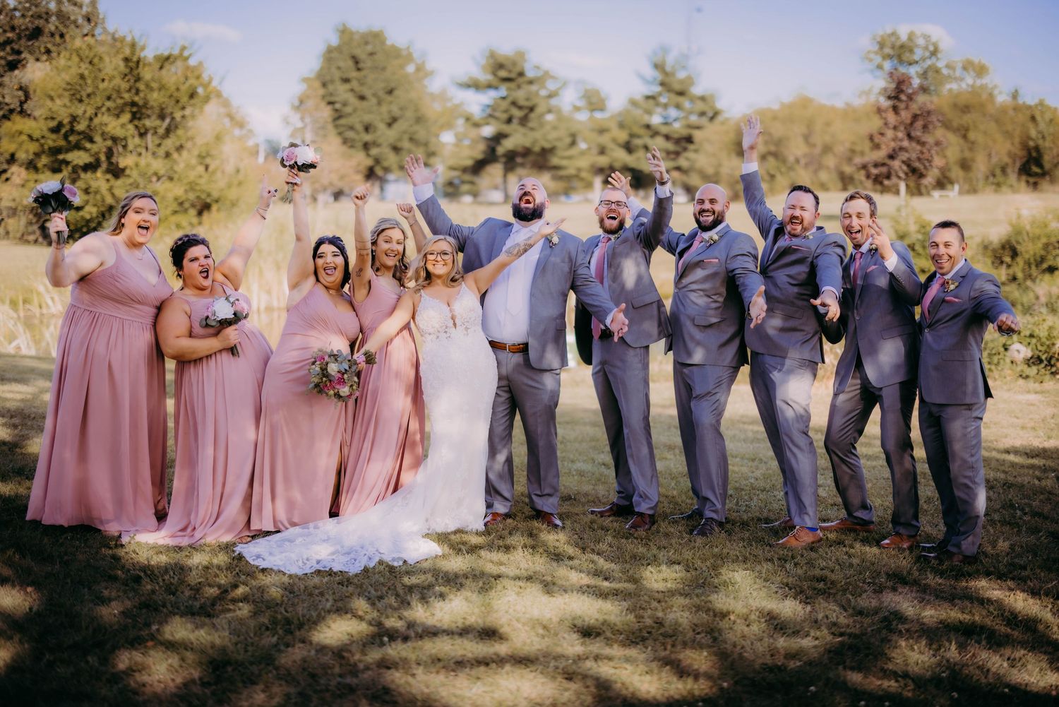 Wedding party in pink dresses and grey suits posing joyfully outdoors.